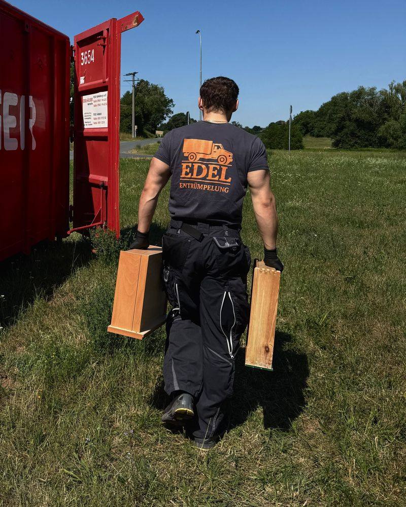 Mitarbeiter von Edel Entrümpelung Ansbach trägt Holzschubladen über eine Wiese zum Container